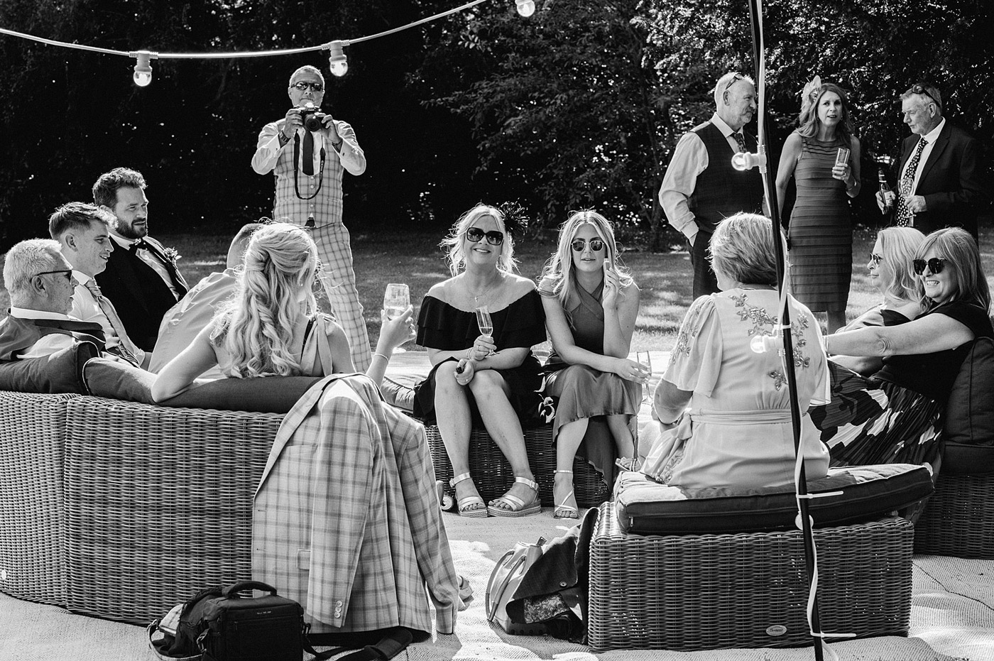 A group of people sit and chat on outdoor wicker furniture at a gathering; some hold drinks, while others stand and talk in the background. One man is photographing the scene. The setting appears relaxed and sociable.