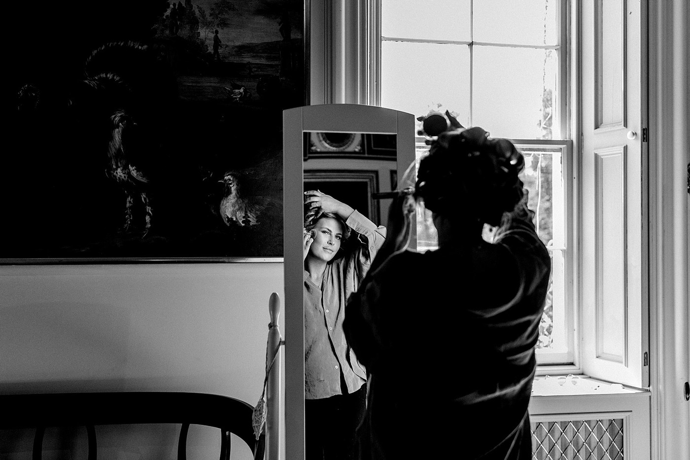 A person stands in front of a mirror, adjusting their hair, while their reflection shows them smiling. The scene is inside a room with a large window and artwork on the wall. The photo is in black and white.