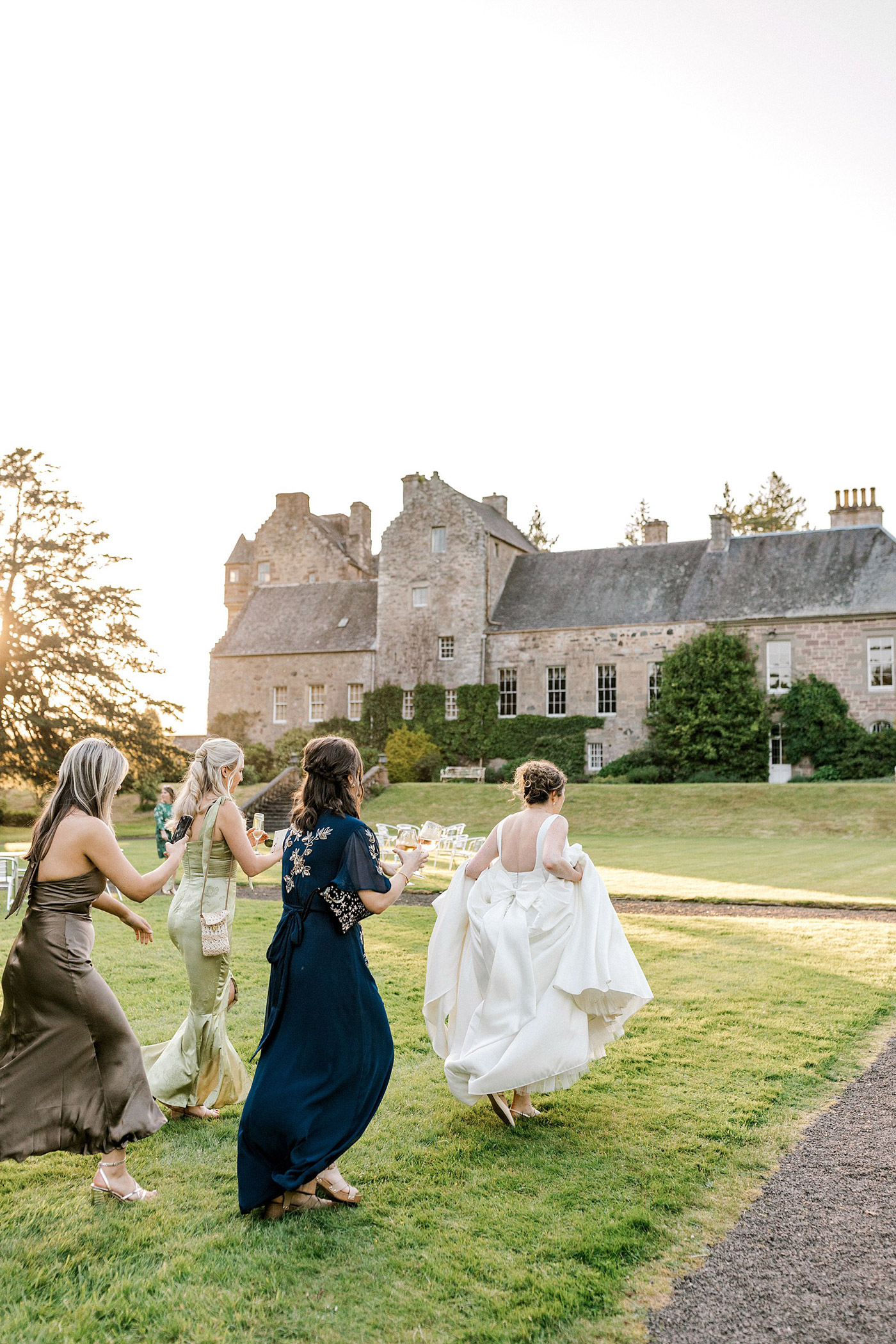 Four women in elegant dresses walk and laugh together on a grassy lawn in front of a large, stone historic building under a bright sky. The bride, holding her dress, leads the group.