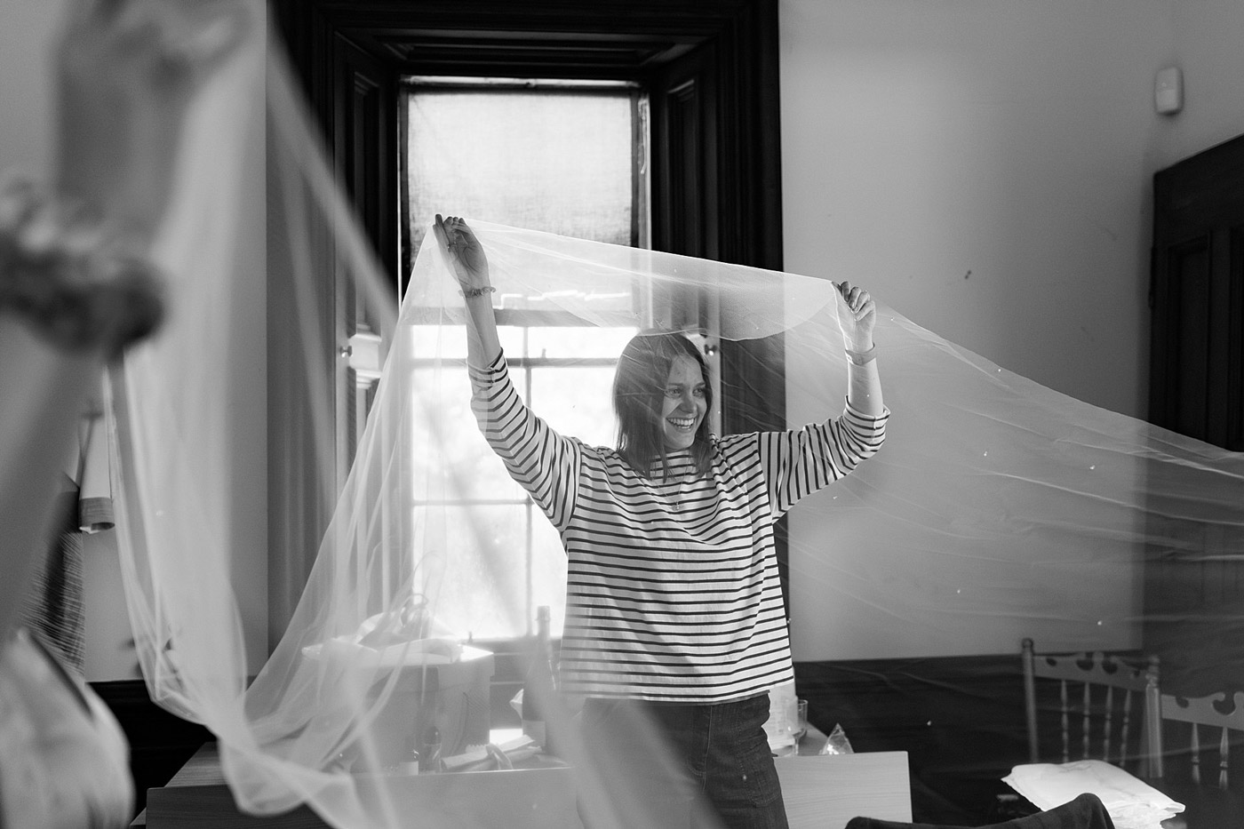 A woman smiling and holding up a long piece of sheer fabric in a room with a large window behind her. The scene is in black and white.