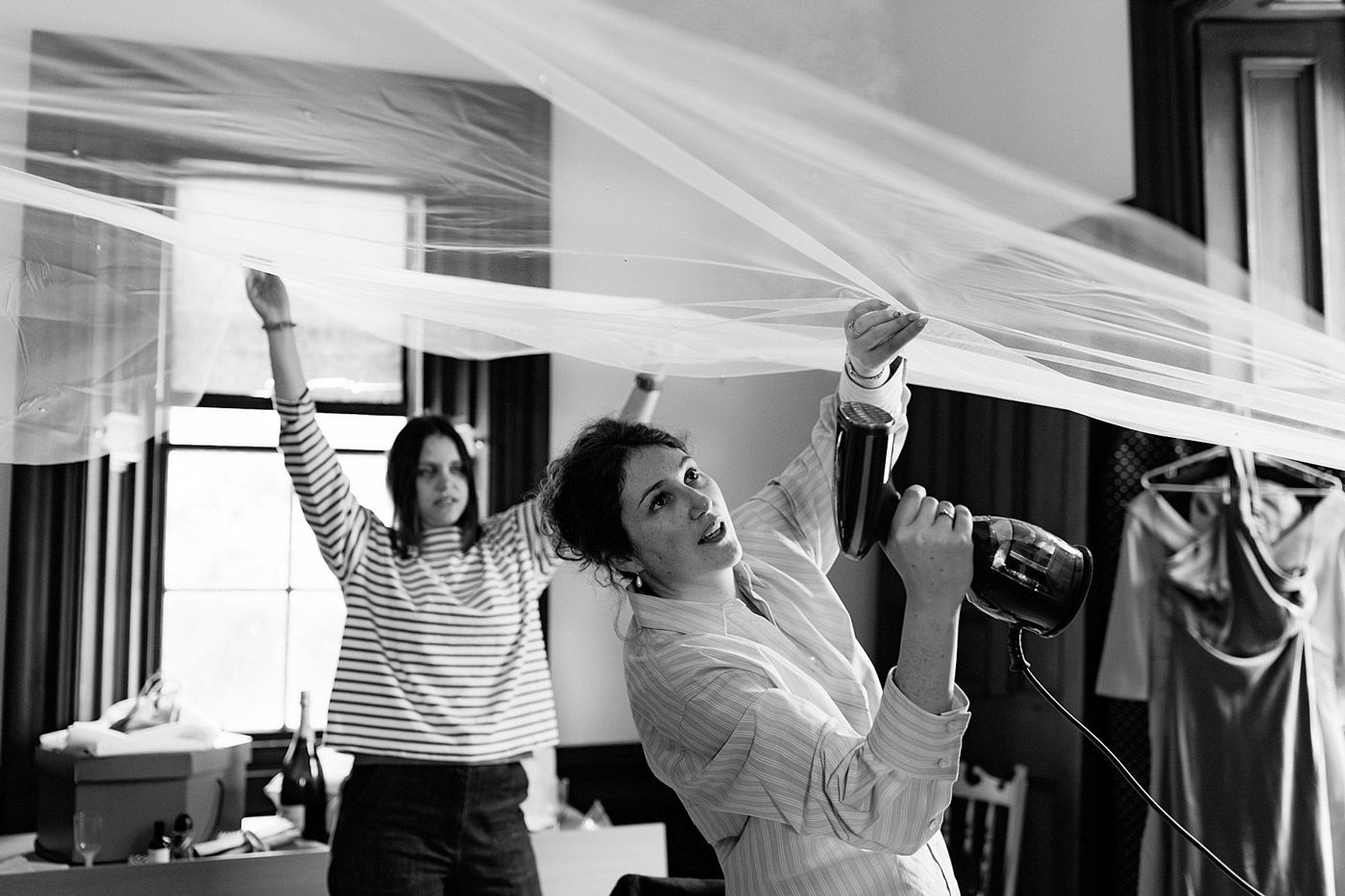 Two women work together indoors to hang a large piece of sheer fabric; one holds it up while the other uses a hairdryer, possibly to smooth or secure the material. The scene is candid and in black and white.
