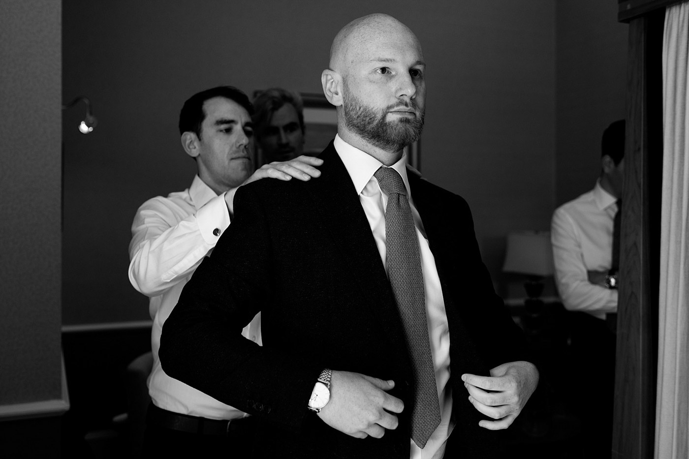 A man in a suit stands confidently while another man behind him adjusts his jacket; two other men in dress shirts are visible in the background, all preparing in a dimly lit room.