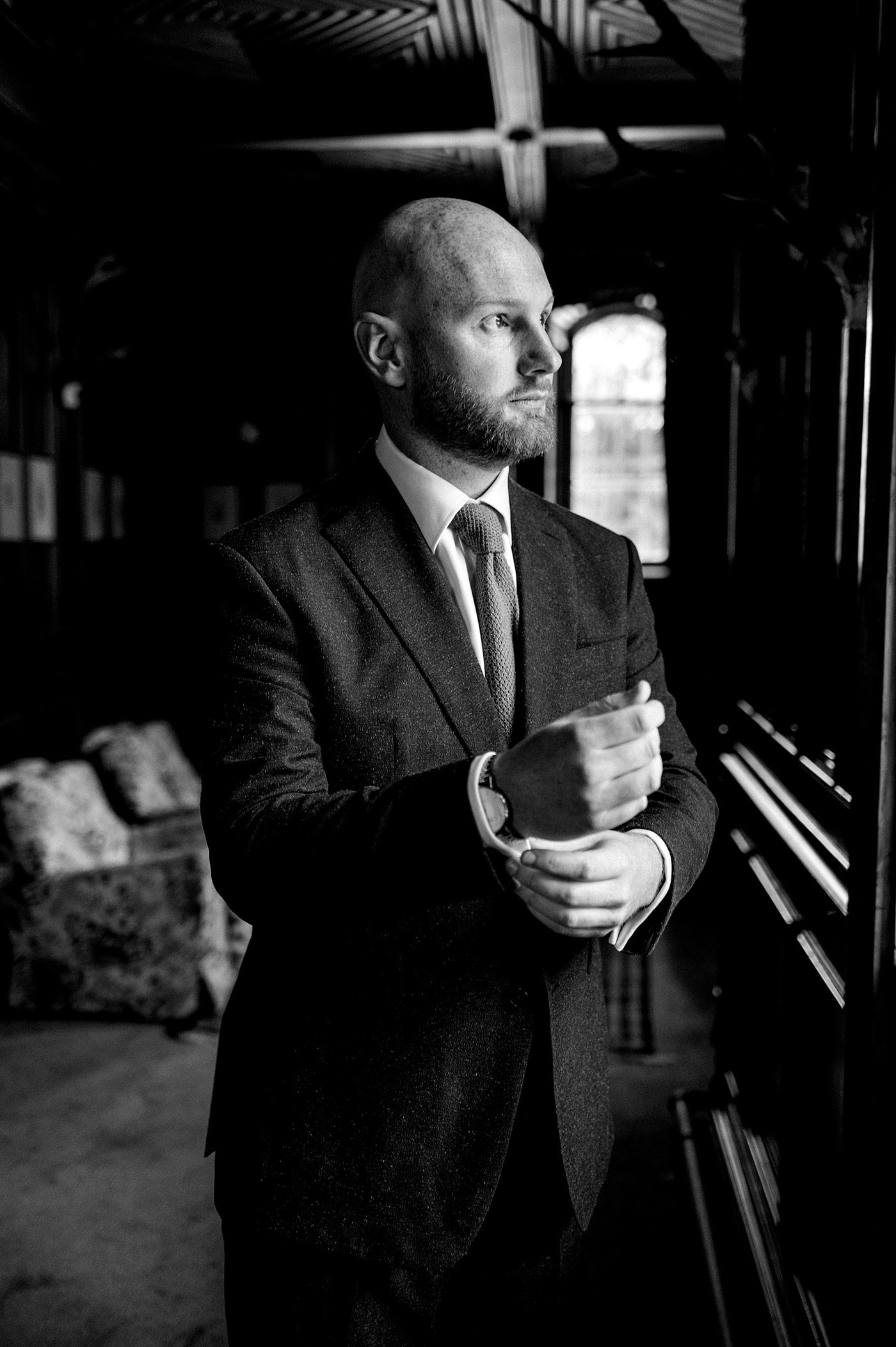 A man in a suit stands indoors near a window, adjusting his cufflink. The black-and-white photo captures him looking thoughtfully into the distance, with light streaming in from the window behind him.