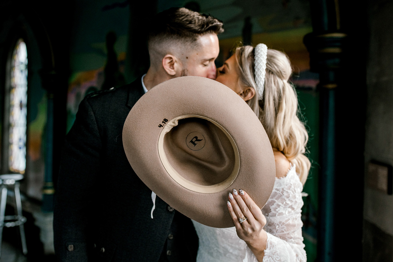 A bride and groom share a kiss while the bride holds a beige hat in front of their faces, partially hiding their kiss. The bride wears a white lace dress and headband, and the groom is in a dark suit.