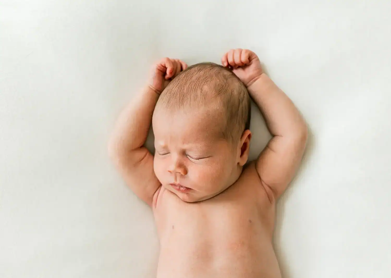 A newborn baby sleeps peacefully on a white blanket with arms raised above their head and eyes closed.