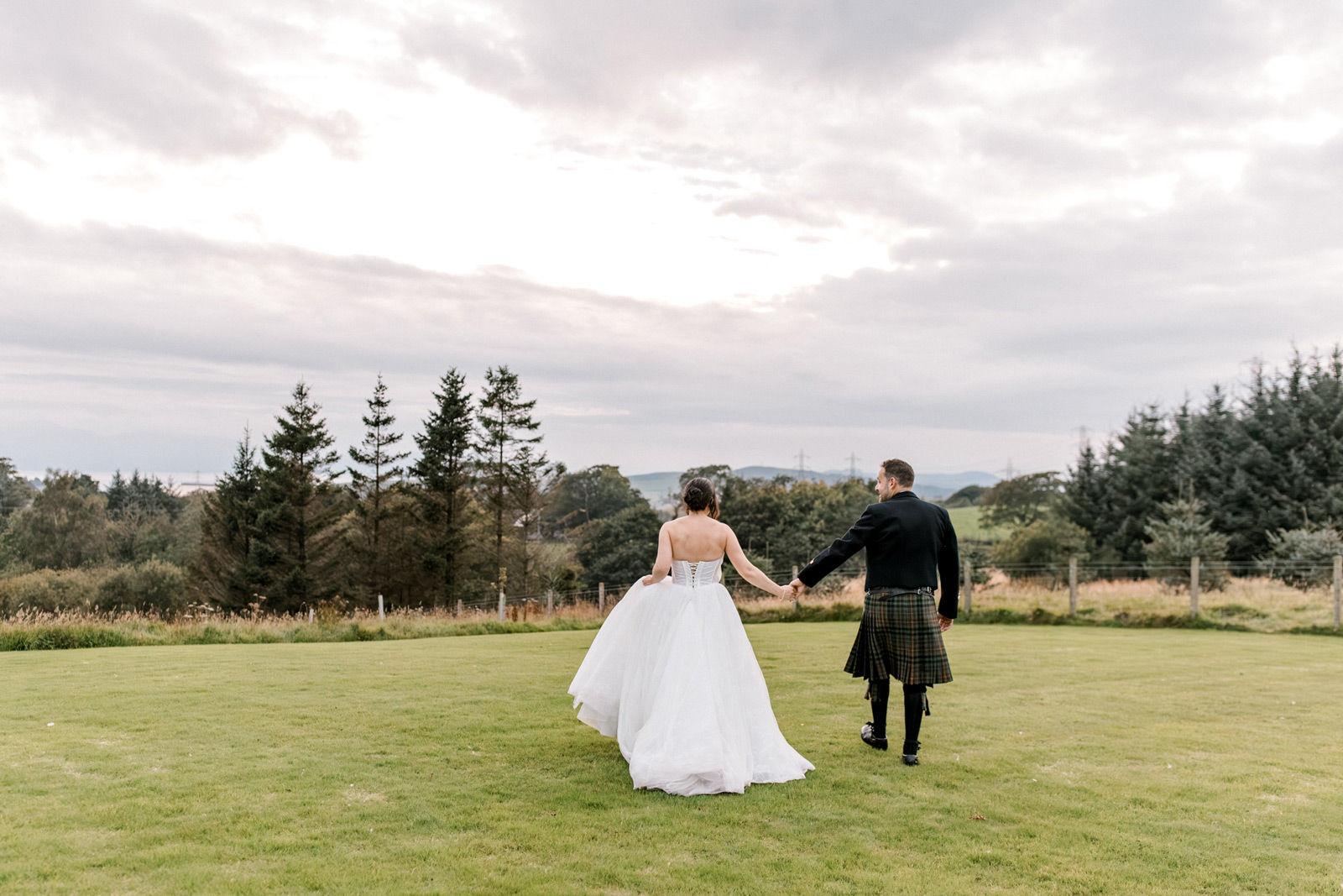 A bride in a white gown and a groom in traditional Scottish attire walk hand in hand across a grassy field, with trees and cloudy sky in the background.