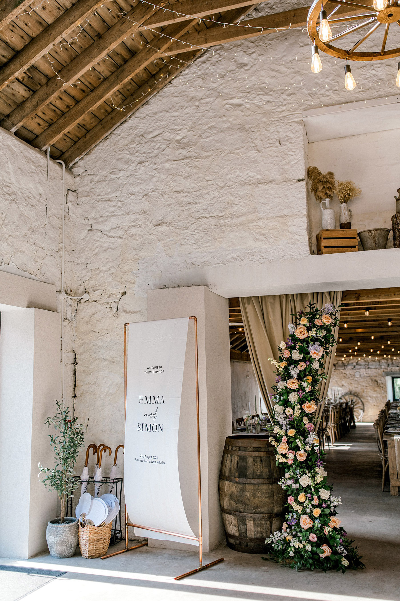 A rustic wedding venue features a welcome sign for Emma and Simon, floral arrangements, a wine barrel, baskets with umbrellas, and fairy lights hanging from a wooden ceiling.