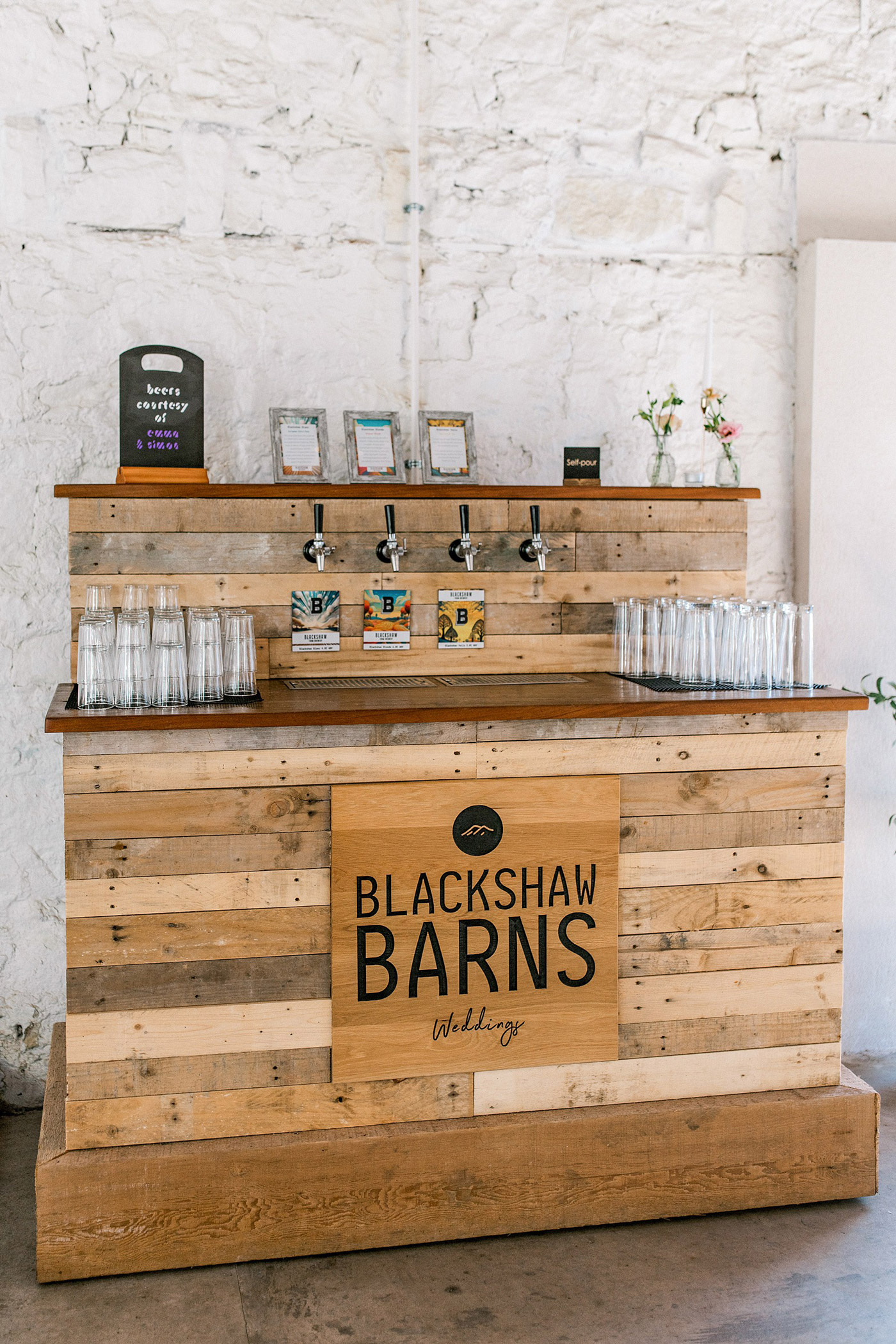 A rustic wooden bar with “Blackshaw Barns Weddings” sign, four beer taps, glassware neatly arranged, and small framed signs on the counter, set against a white stone wall.