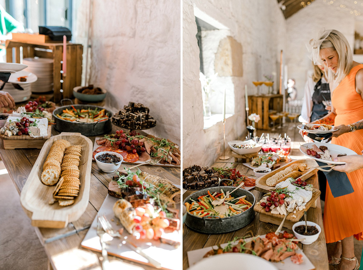 A rustic buffet table is set with assorted cheeses, biscuits, fruits, meats, and desserts. On the right, a woman in an orange dress helps herself to food from the spread inside a bright, stone-walled room.