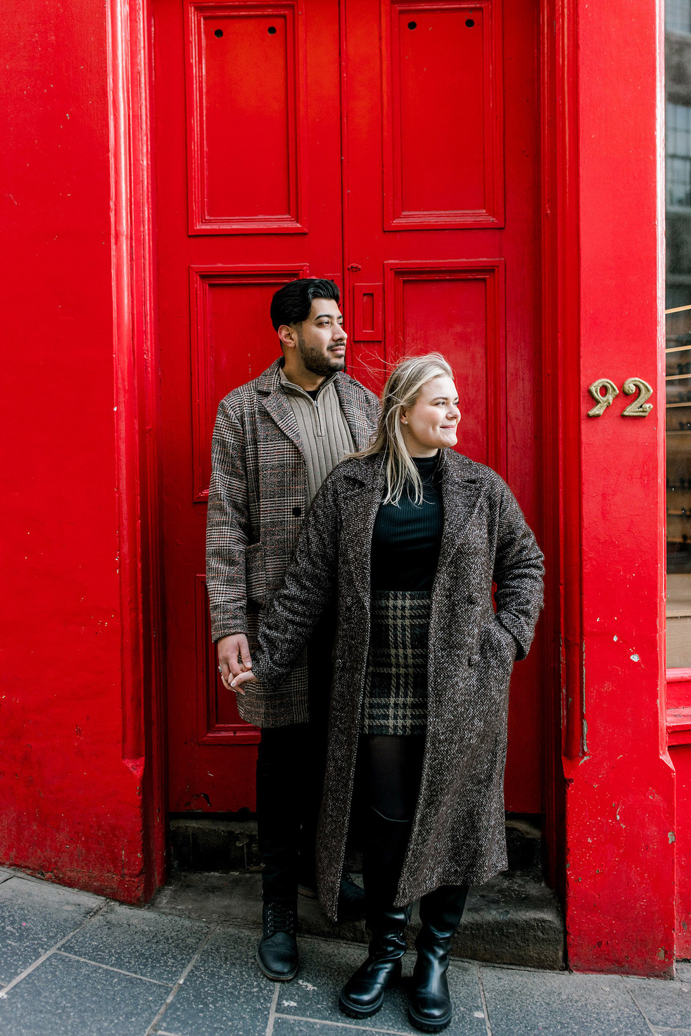 A couple stands holding hands in front of a bright red door numbered 92. Both wear long winter coats and boots. The woman looks to the side, whilst the man looks ahead, standing closely behind her.