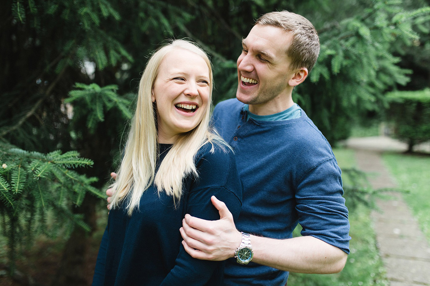 A smiling couple stands close together outdoors near green pine trees. Both people are wearing blue tops, laughing and looking happy, with the man's arm around the woman's shoulder.