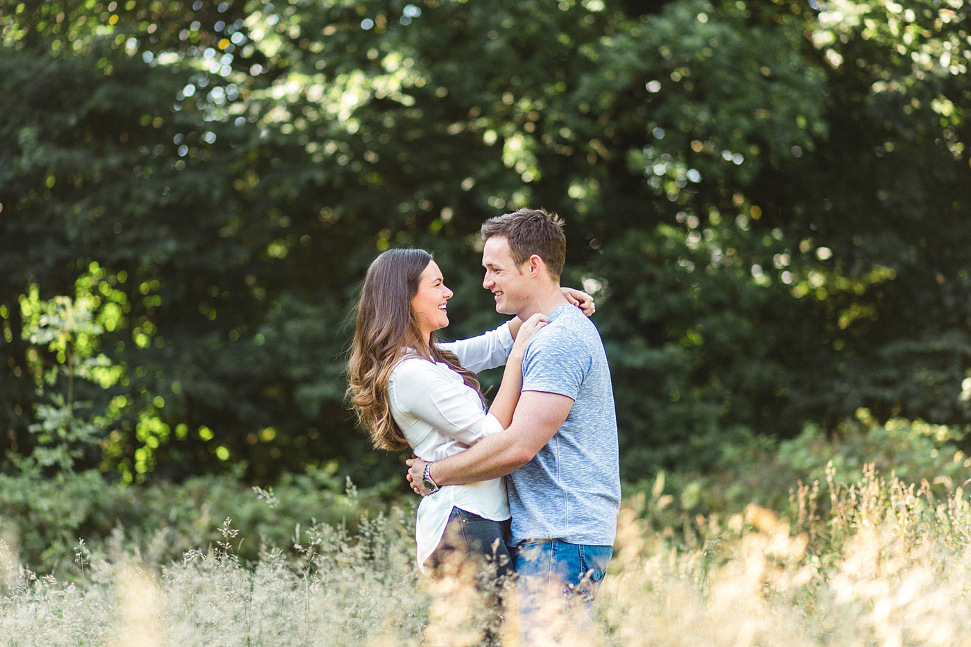 A couple stands in a grassy field, holding each other and smiling, surrounded by green trees in the background on a sunny day.