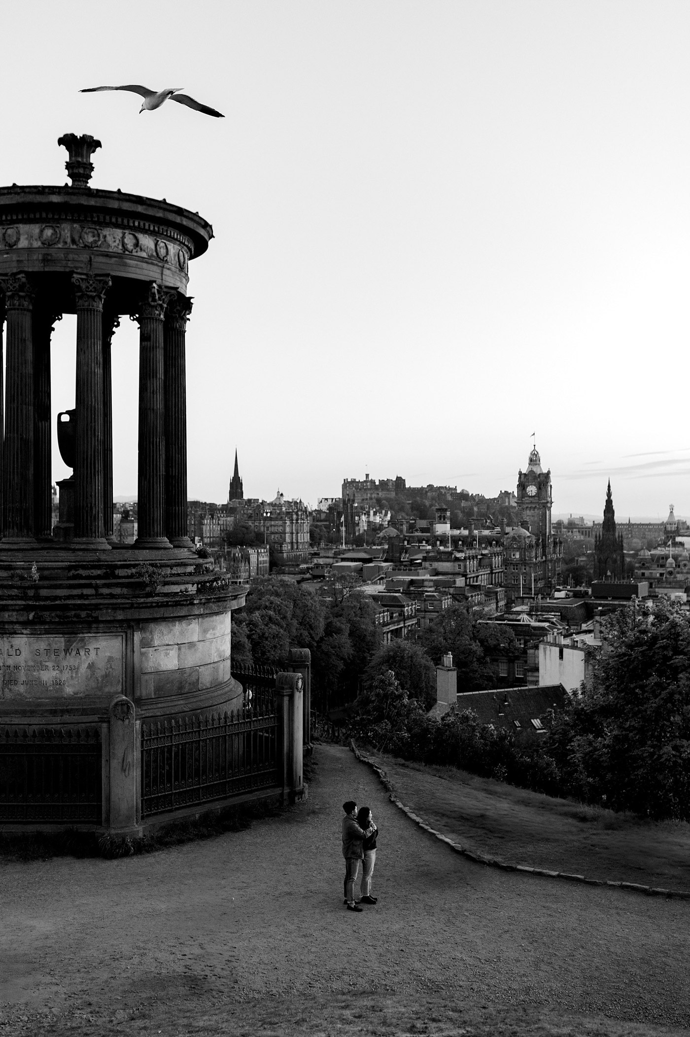 A black-and-white photo of a couple standing near the Dugald Stewart Monument, overlooking Edinburgh’s cityscape with a flying seagull in the sky and historic buildings in the distance.