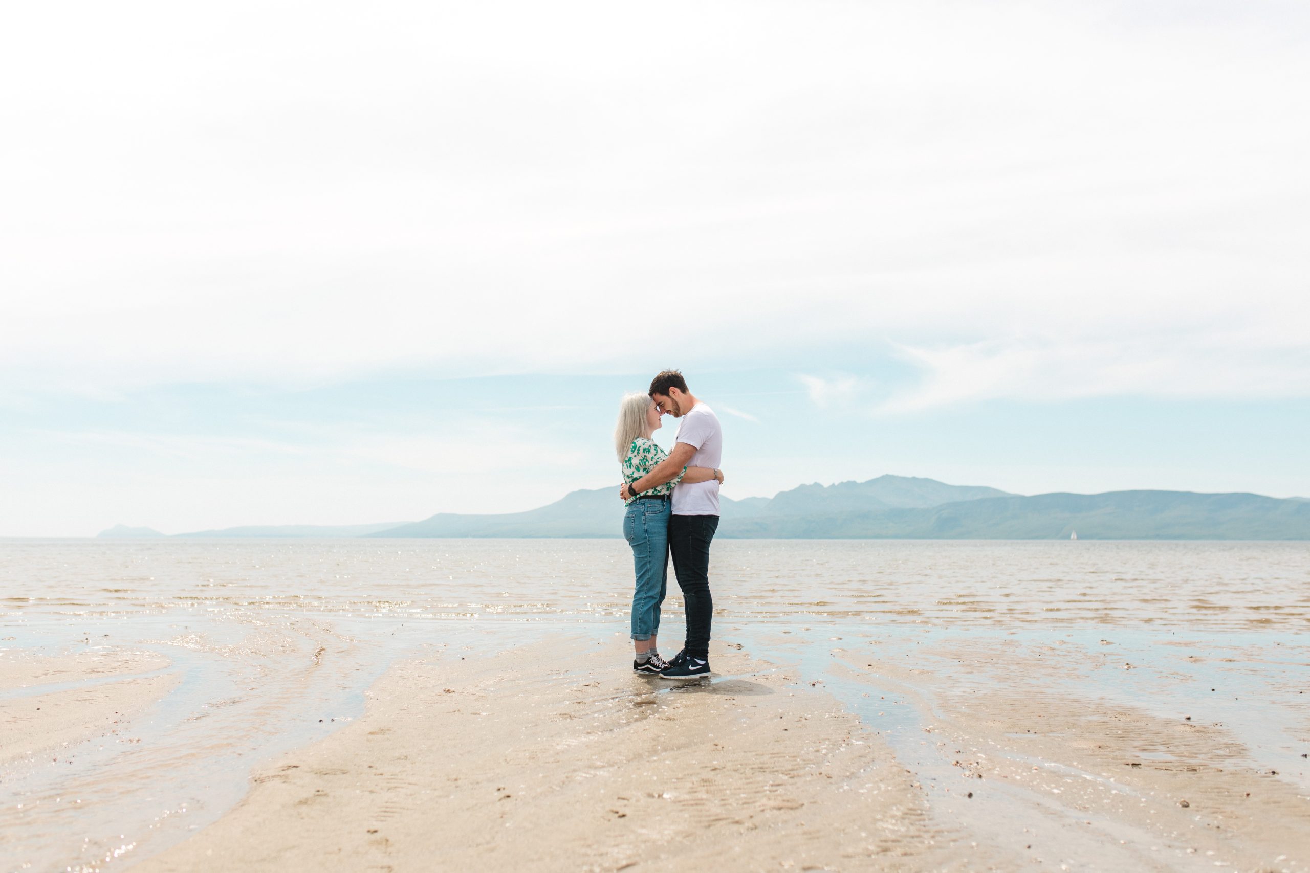 A couple stands close together on a sandy beach, embracing and touching foreheads, with mountains and a calm sea in the background under a bright sky.