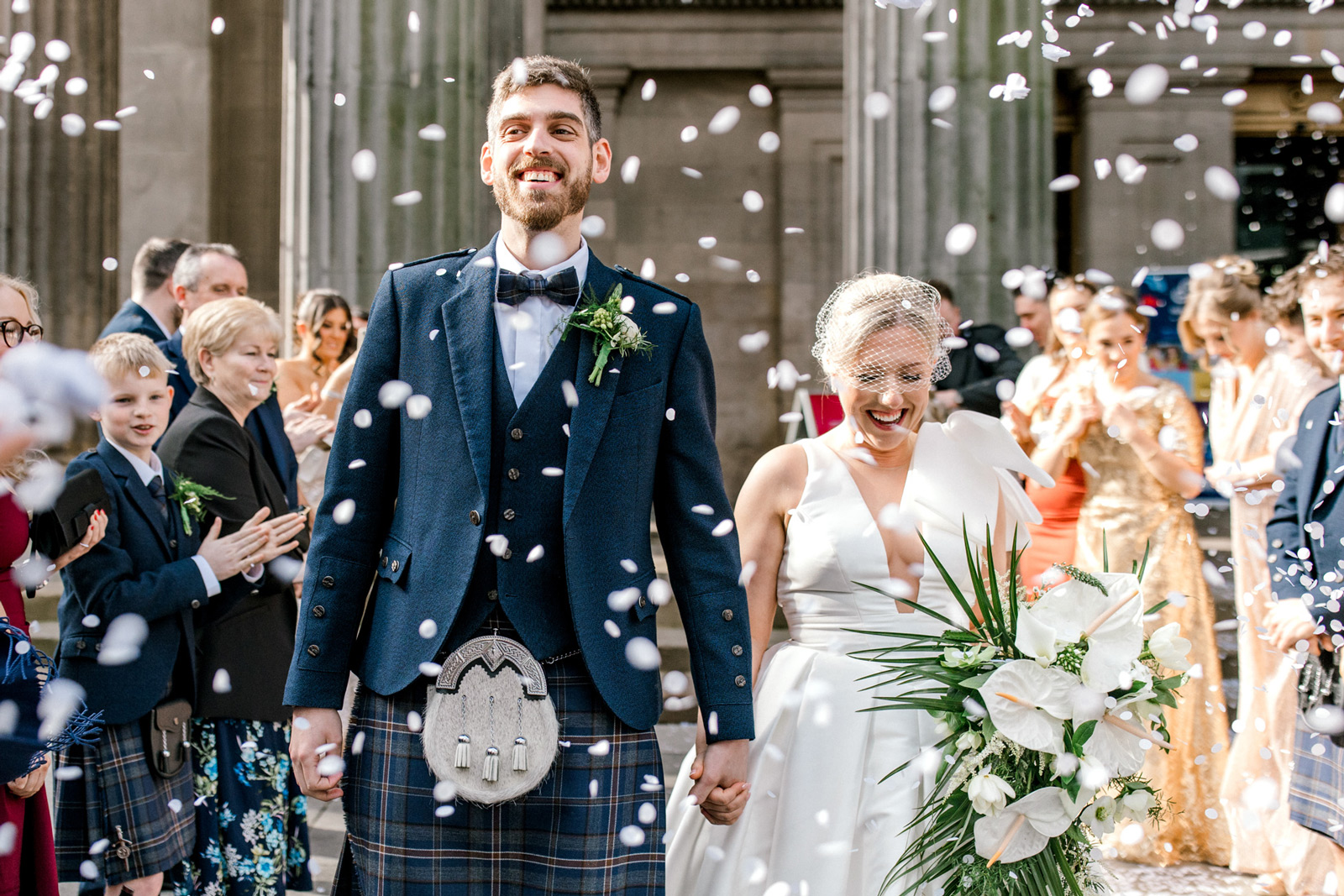 A smiling bride and groom walk hand-in-hand outdoors whilst guests throw confetti. The groom wears a kilt and jacket; the bride wears a white dress and holds a bouquet of white flowers. Guests clap and celebrate in the background.