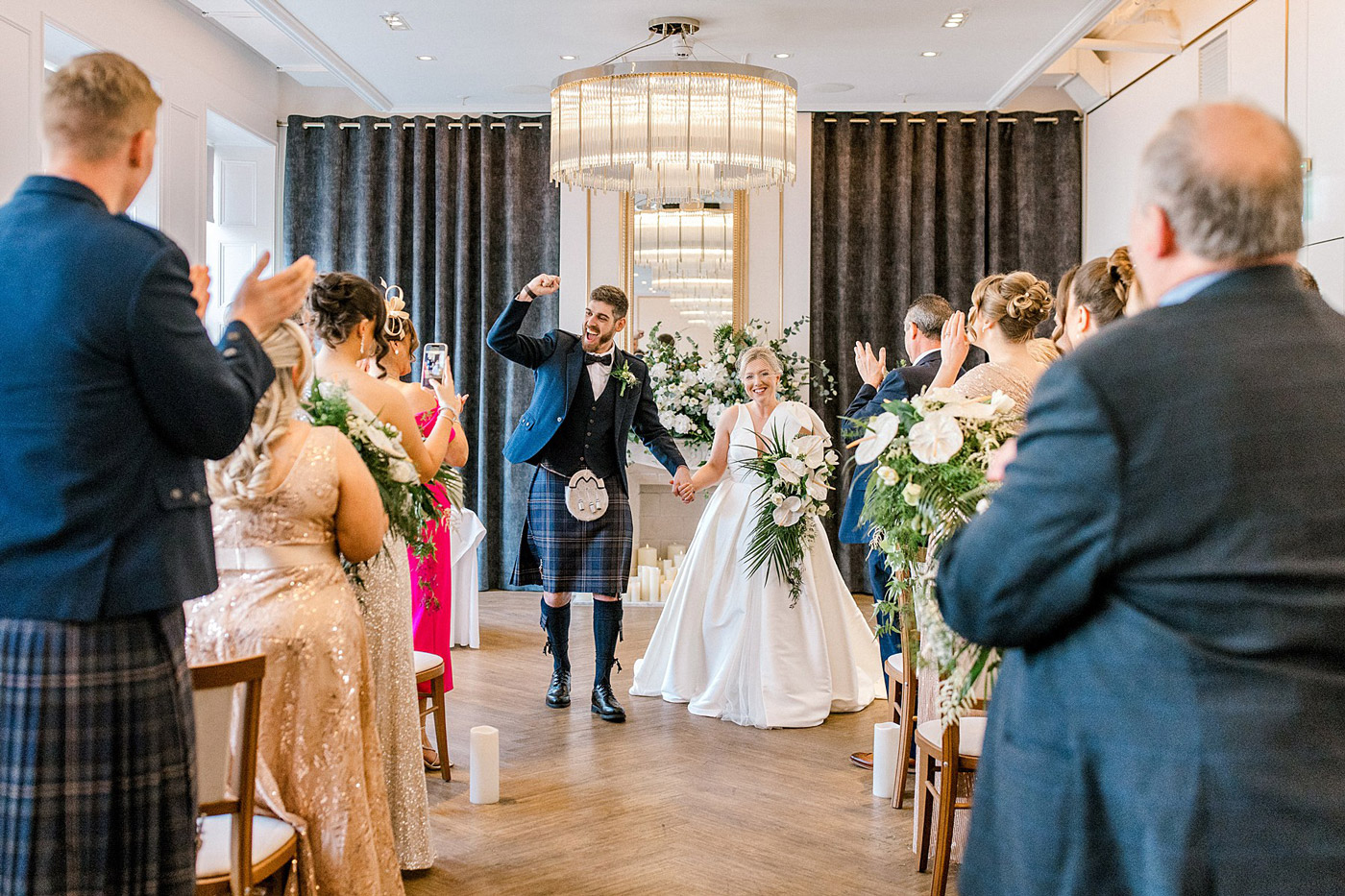 A joyful bride and groom walk hand in hand down the aisle as guests clap and celebrate. The groom raises his fist in excitement, and the elegant indoor venue is decorated with flowers and a large chandelier.