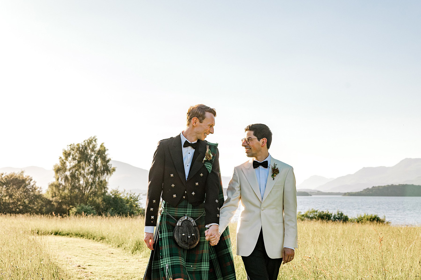 Two men in wedding attire, one in a traditional Scottish kilt and the other in a white dinner jacket, walk hand in hand, smiling at each other in a sunlit field with mountains and a lake in the background.