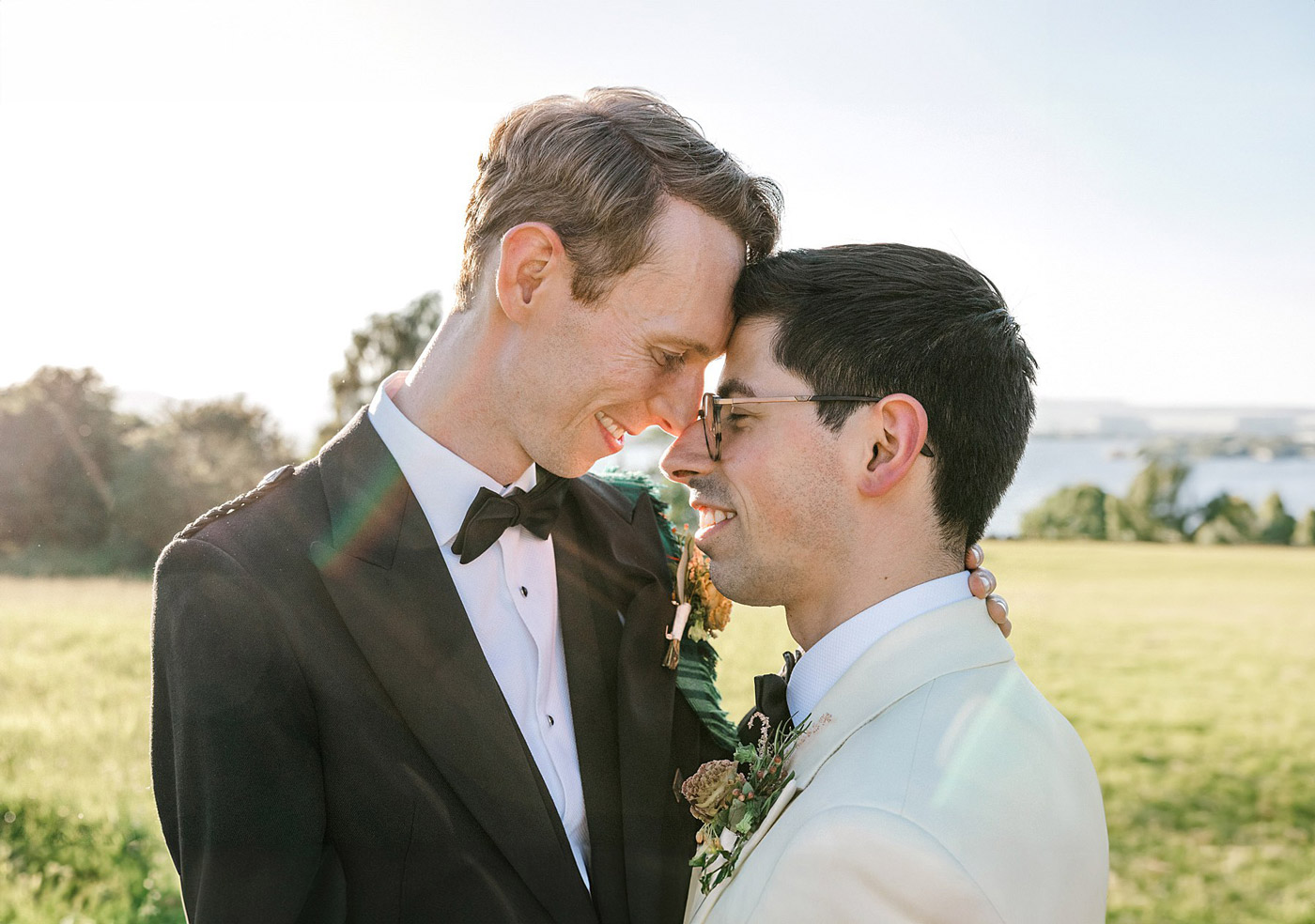 Two grooms in tuxedos stand closely together outdoors, smiling and touching foreheads lovingly, with a field and water in the background, bathed in warm sunlight.