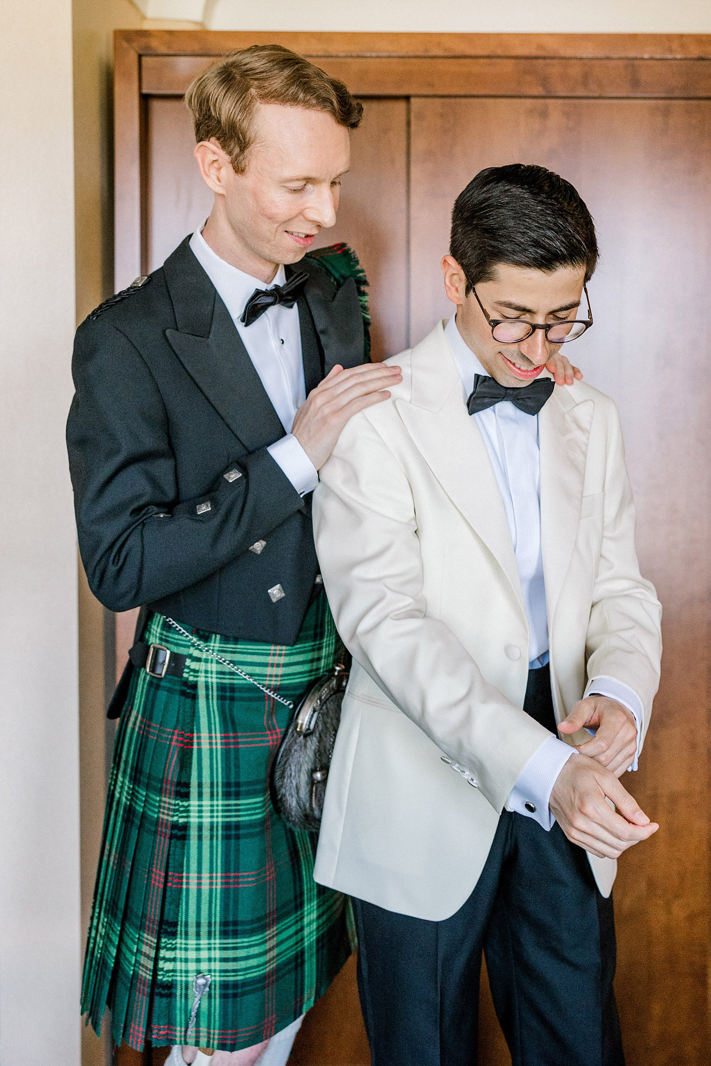 Two men dressed formally stand indoors; one wears a black jacket and green tartan kilt, gently touching the other’s shoulder as he puts on a white dinner jacket and adjusts his cufflinks, both smiling.