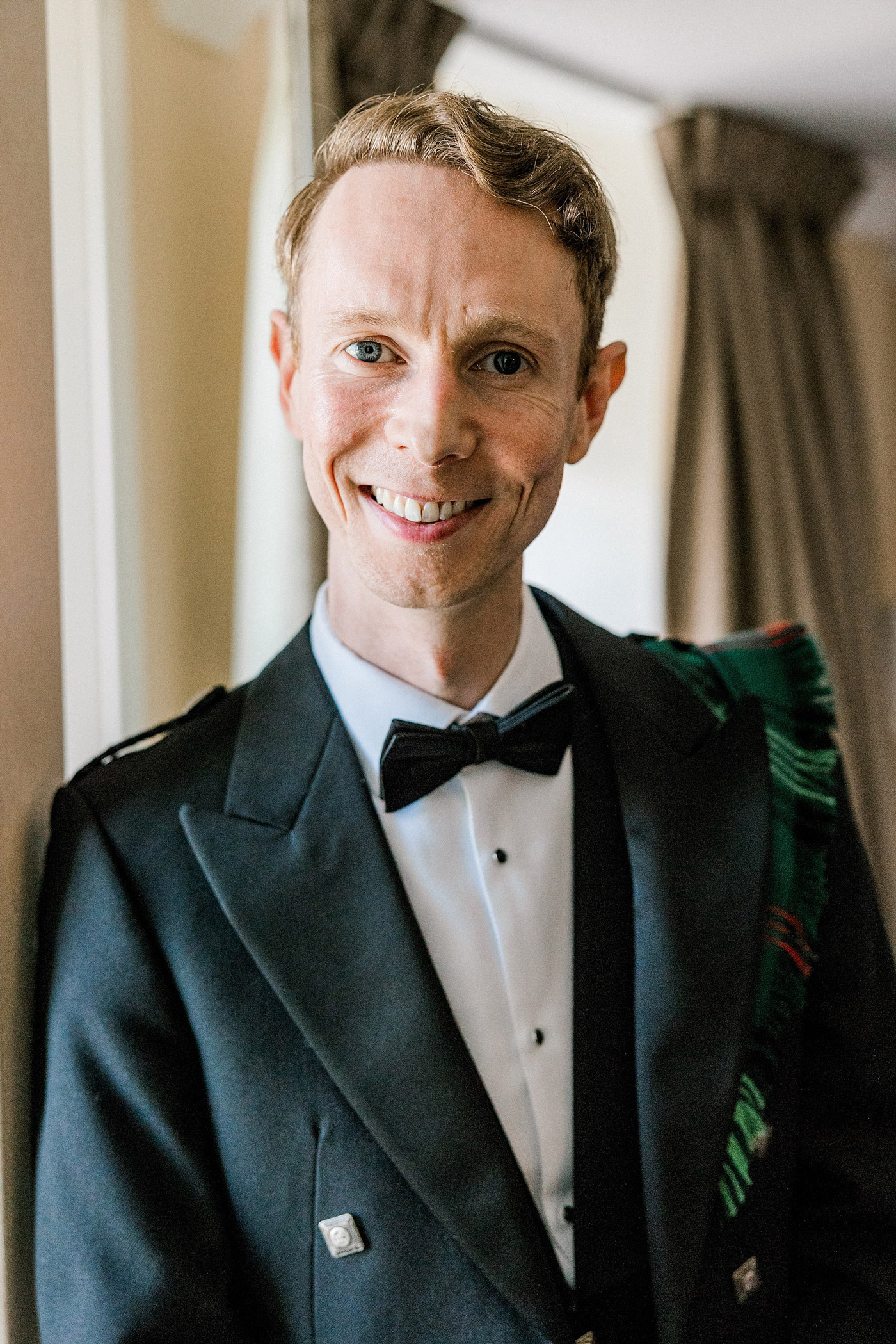 A smiling man with short light brown hair wears a black dinner jacket with a bow tie and a tartan sash, standing indoors near a window with beige curtains in the background.