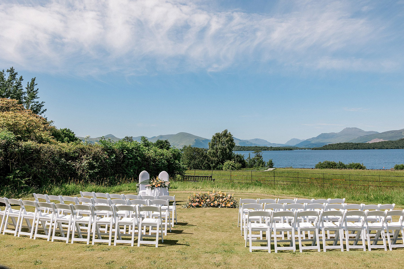Rows of white chairs set up on a grassy lawn for an outdoor wedding ceremony, facing two decorated chairs and floral arrangements, with a scenic lake, mountains, and blue sky in the background.
