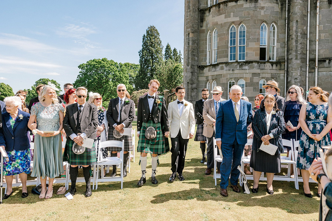 A wedding ceremony outdoors with guests standing in rows. Two men, one in a white tuxedo and one in a black jacket and kilt, walk together down the aisle. The backdrop is a large stone building and sunny blue sky.