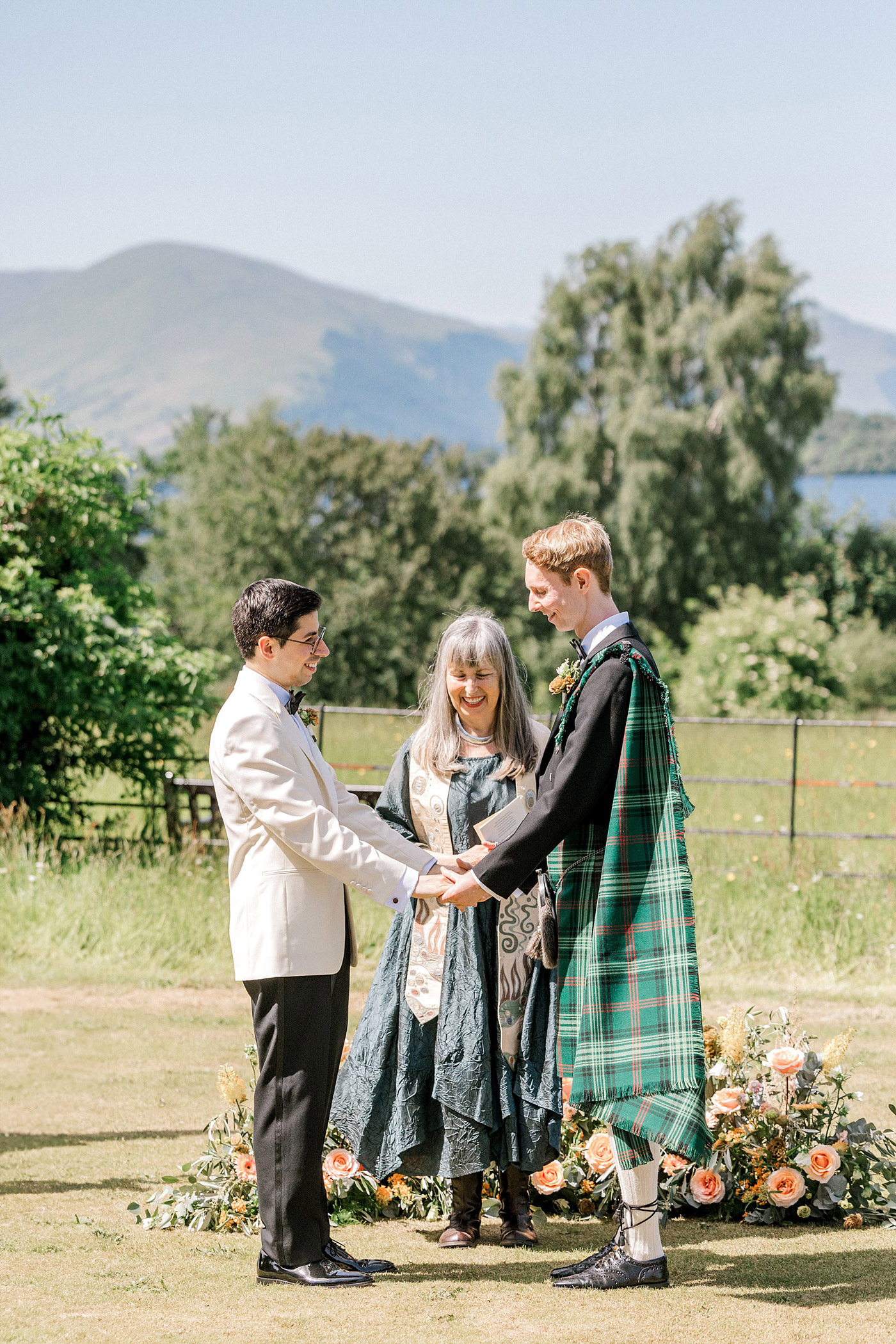 Two grooms stand holding hands in front of an officiant outdoors, surrounded by flowers and greenery, with mountains and trees in the background under a clear sky.