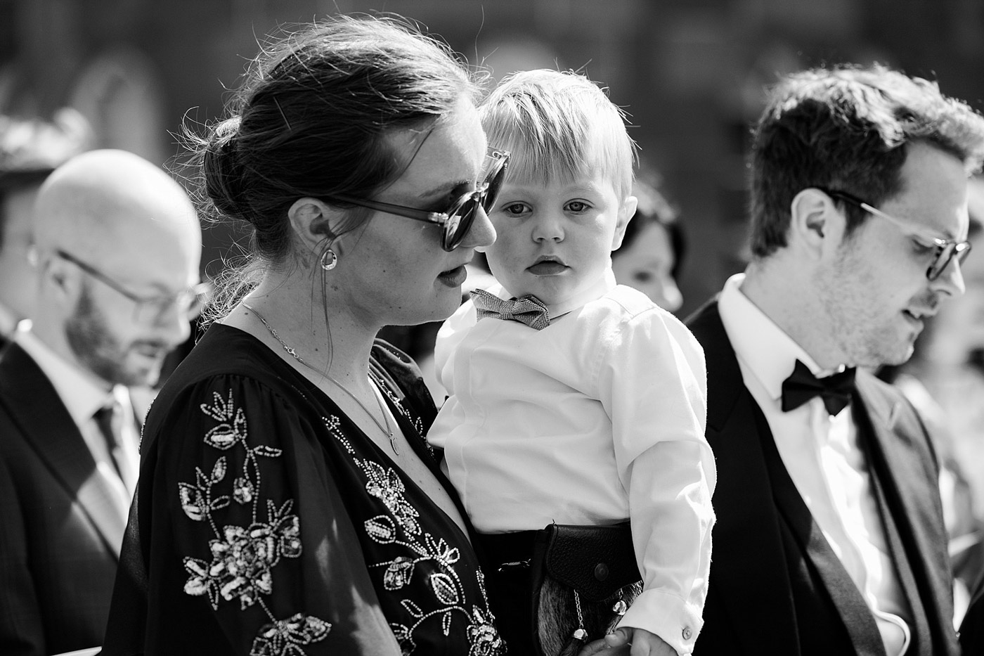 A woman wearing sunglasses holds a young child dressed in a bow tie and waistcoat at an outdoor event, surrounded by people in formal attire. The image is in black and white.