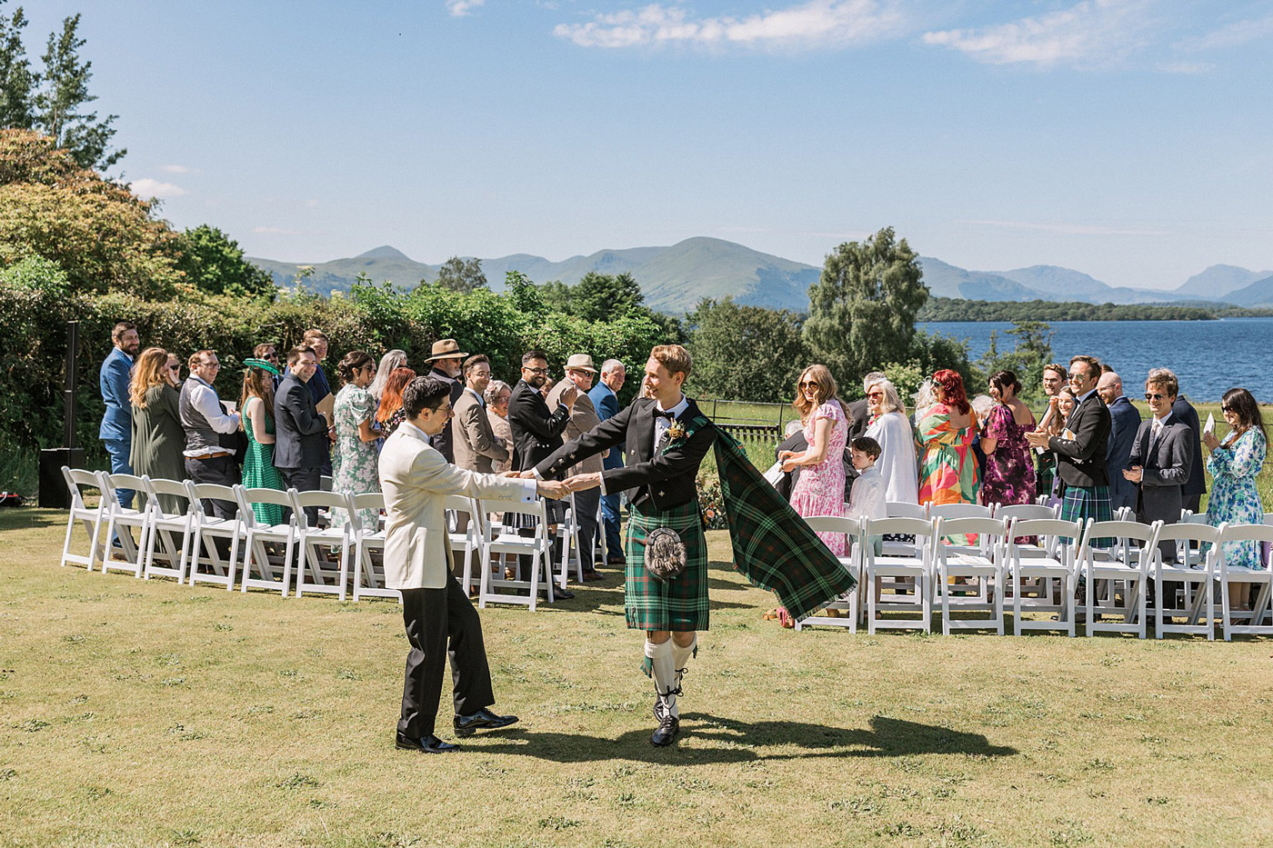 Two grooms, one in a white jacket and the other in a traditional Scottish kilt, walk hand in hand down an outdoor aisle as guests stand and clap, with a lake and mountains in the background.
