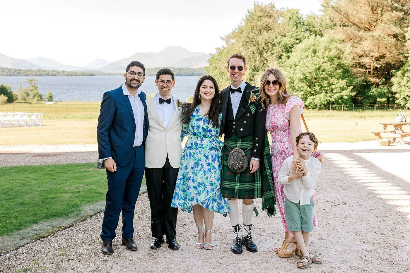Six people, including one wearing a kilt and another in a blue suit, stand smiling together outdoors on a sunny day, with a lake, mountains, and greenery in the background. A young boy stands in front, barefoot and smiling.