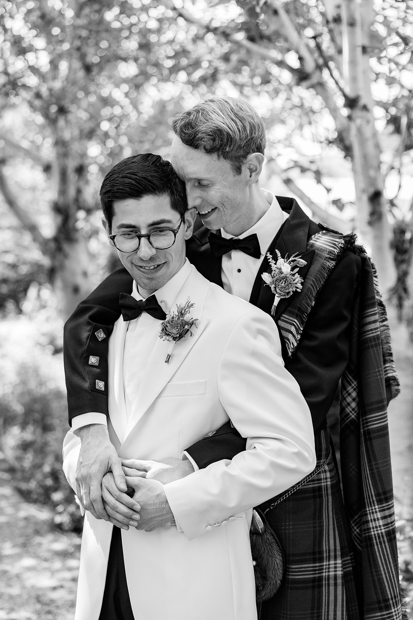 Two grooms stand outdoors, one in a white tuxedo and glasses, the other in a dark suit with a checked sash. The groom in the dark suit hugs the other from behind, both smiling, surrounded by trees.