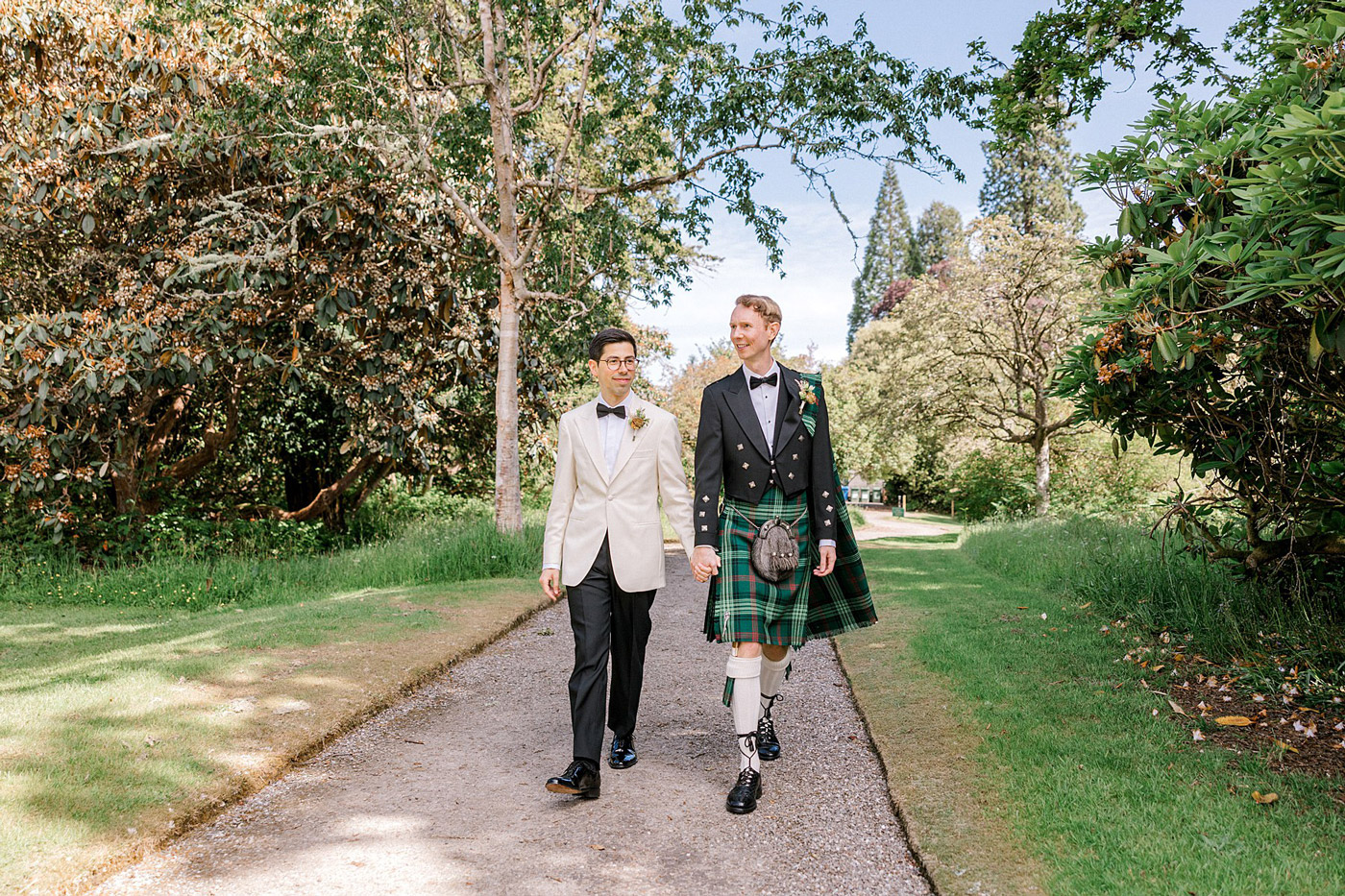 Two men dressed formally, one in a white dinner jacket and the other in traditional Scottish kilt attire, walk hand in hand down a path surrounded by lush greenery and trees on a sunny day.