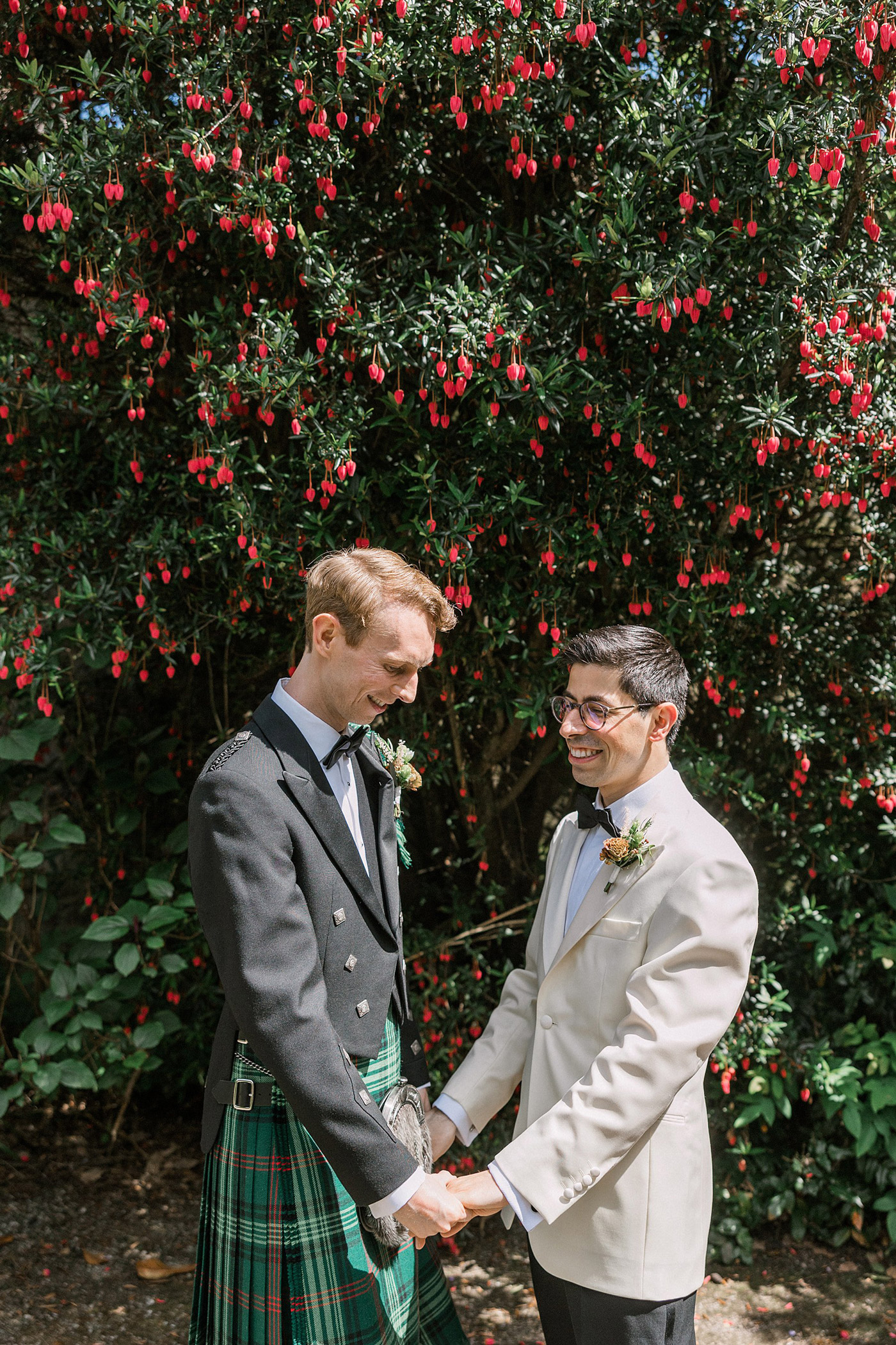 Two grooms stand holding hands and smiling at each other in front of a lush green bush with red flowers. One groom wears a traditional Scottish kilt outfit, and the other wears a white suit jacket with a bow tie.