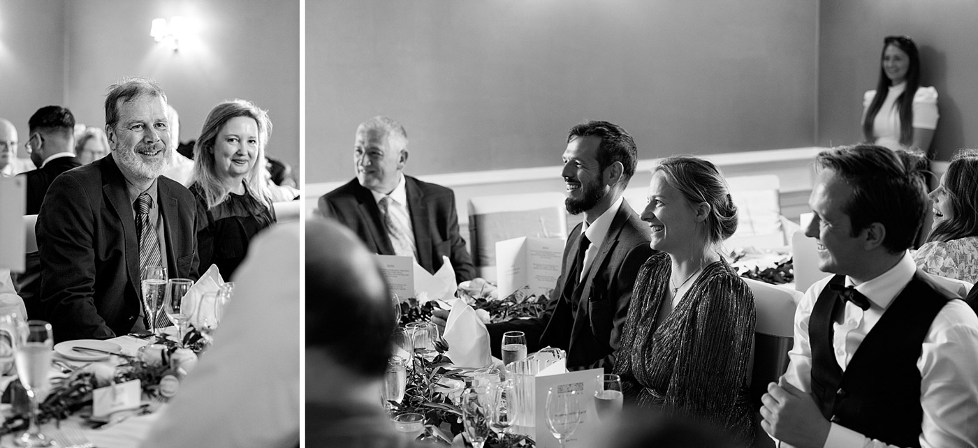 Black and white photo of people sitting at decorated tables, dressed formally, smiling and watching something off camera at what appears to be a celebratory event or reception. A woman stands in the background, also smiling.