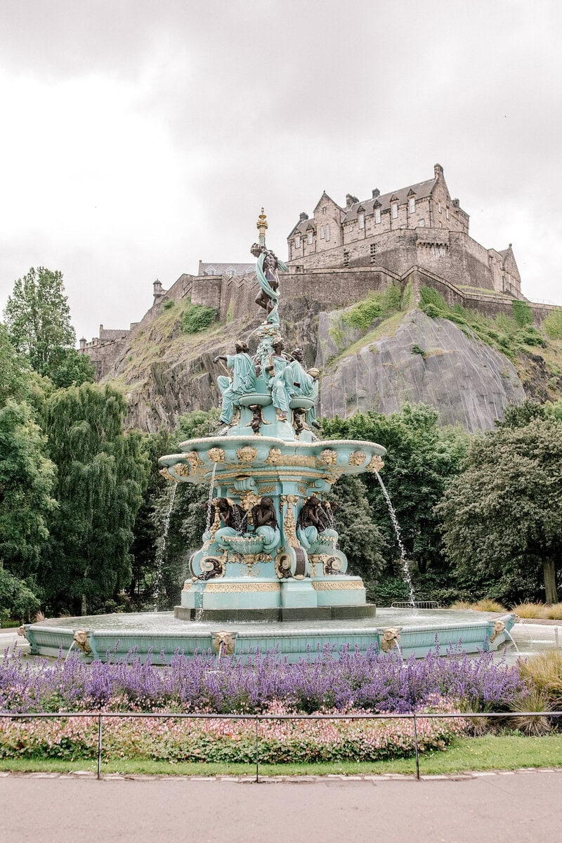 A decorative turquoise fountain with statues is surrounded by flowers and greenery, with Edinburgh Castle perched on a rocky hill in the background under a cloudy sky.