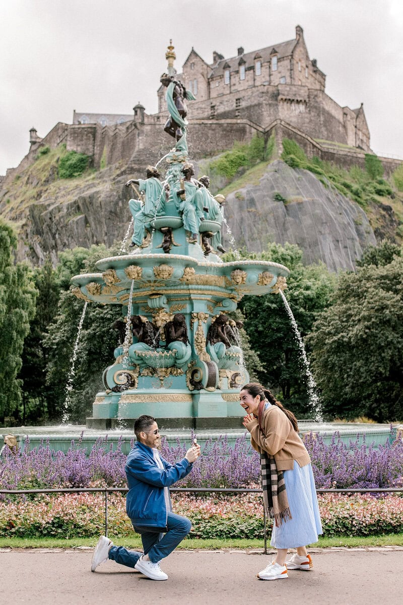A man kneels and proposes to a surprised woman in front of a decorative fountain, with a historic castle on a hill in the background. Gardens with flowers and greenery surround them.