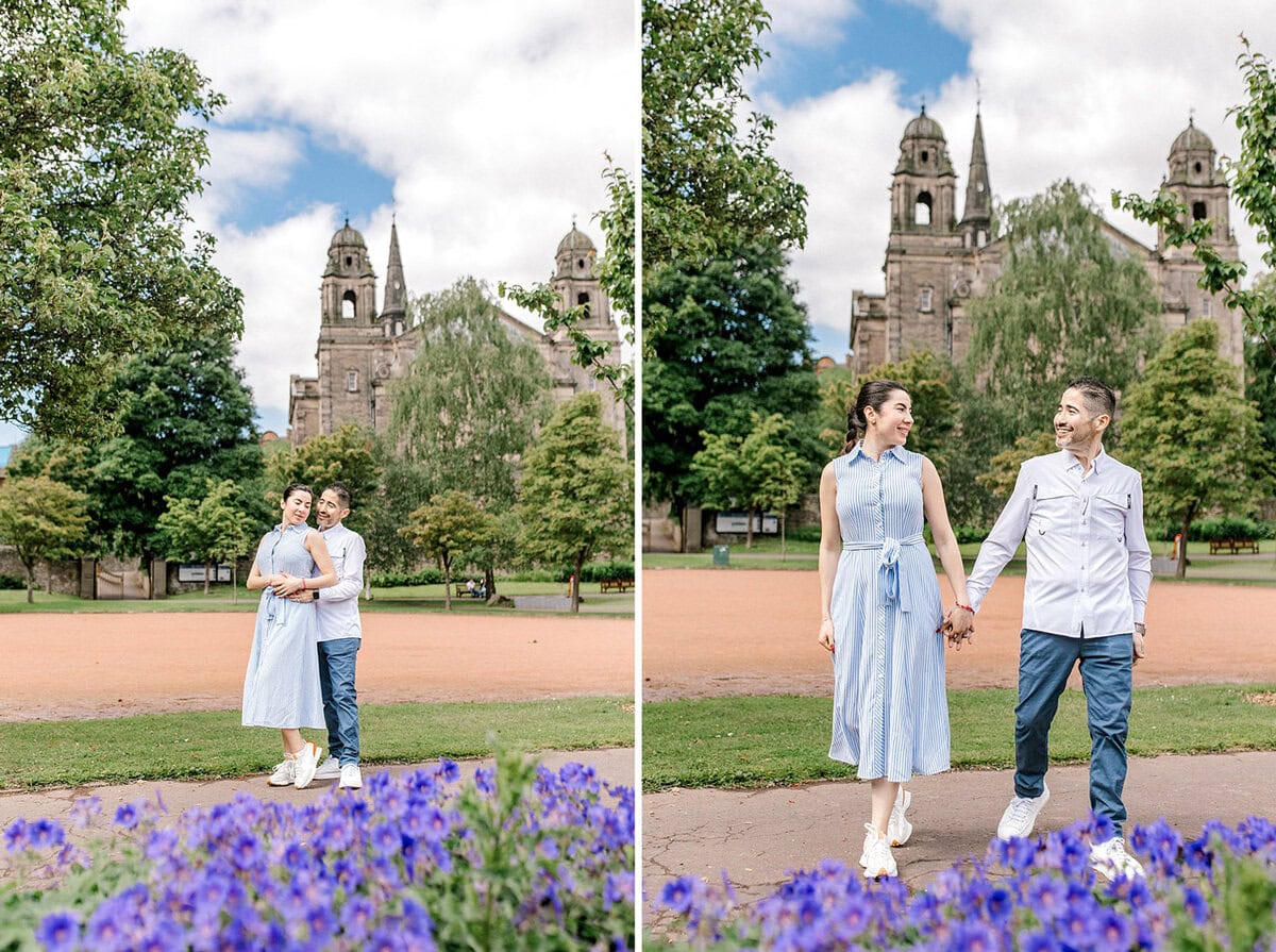 A couple poses and walks together in a park with blooming purple flowers, lush green trees, and a historic stone building with towers in the background under a partly cloudy sky.