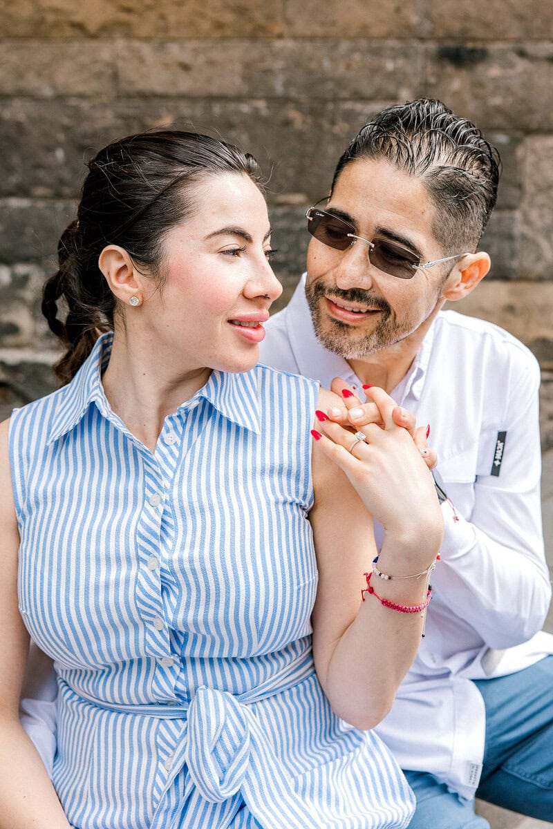 A couple sits closely together. The woman in a blue and white striped sleeveless top smiles whilst showing her ring. The man, wearing glasses and a white shirt, sits behind her, smiling affectionately. A stone wall is in the background.