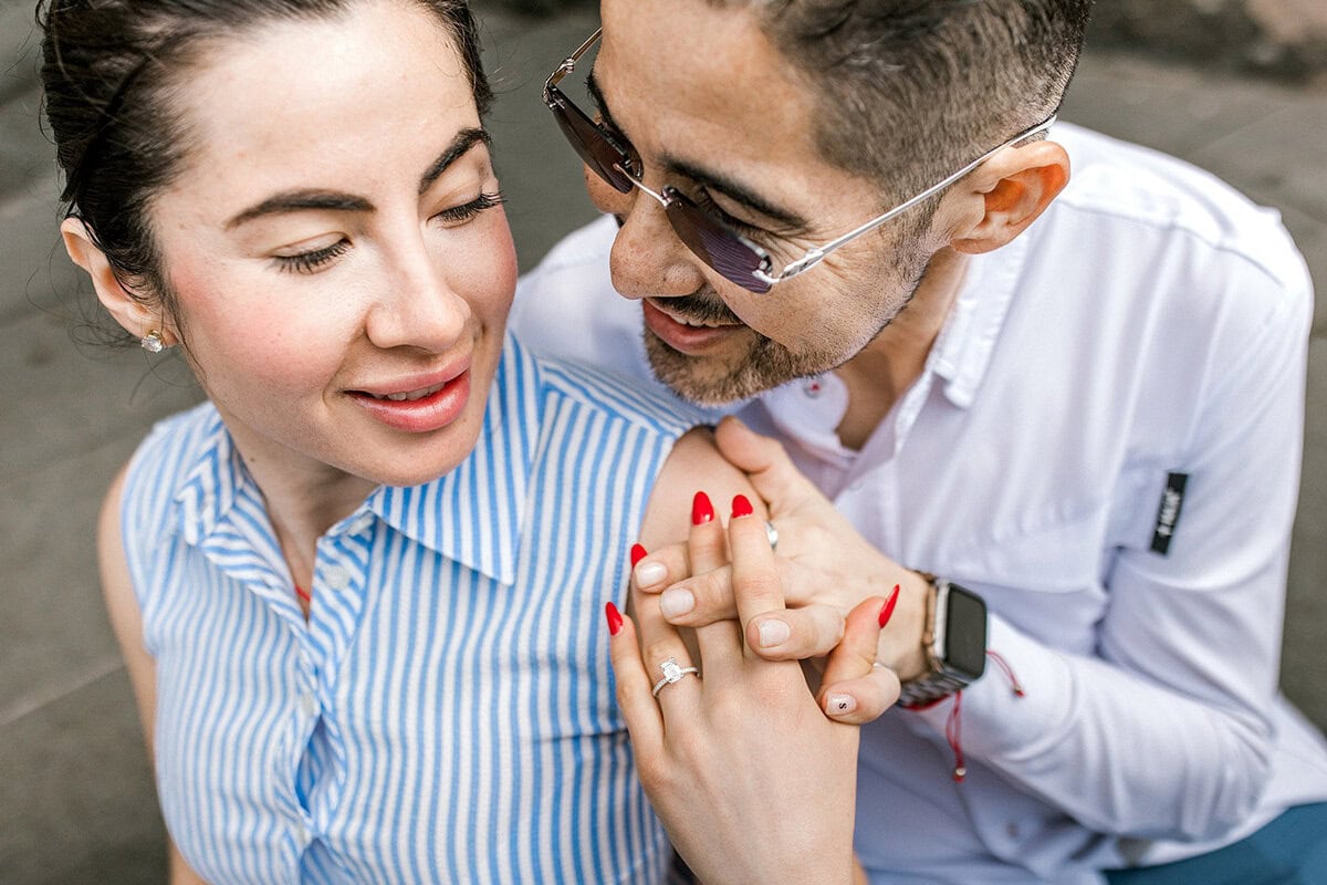 A couple sit close together, smiling warmly. The woman, wearing a blue striped top, shows off an engagement ring as the man, in glasses and a white shirt, lovingly holds her hand.