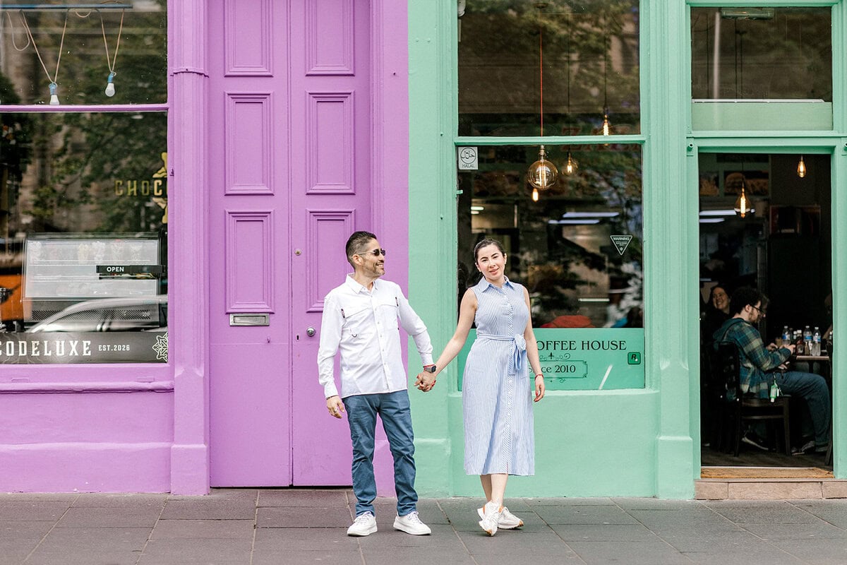 A man and woman hold hands and smile outside colourful shopfronts with purple and green doors and windows. The woman wears a light blue dress; the man wears a white shirt and jeans. People sit inside the café behind them.