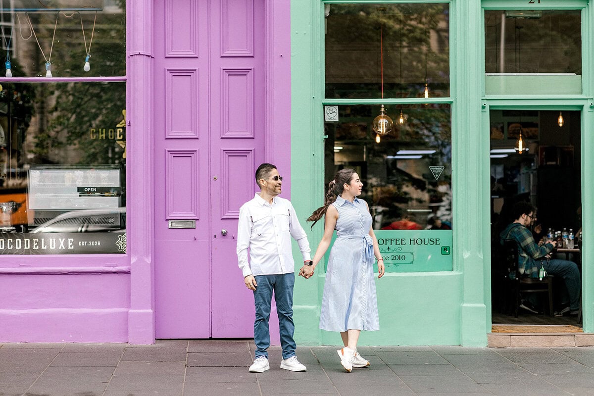A couple hold hands while standing on a pavement in front of colourful purple and green shopfronts, looking in opposite directions. People are seated inside the coffee house.