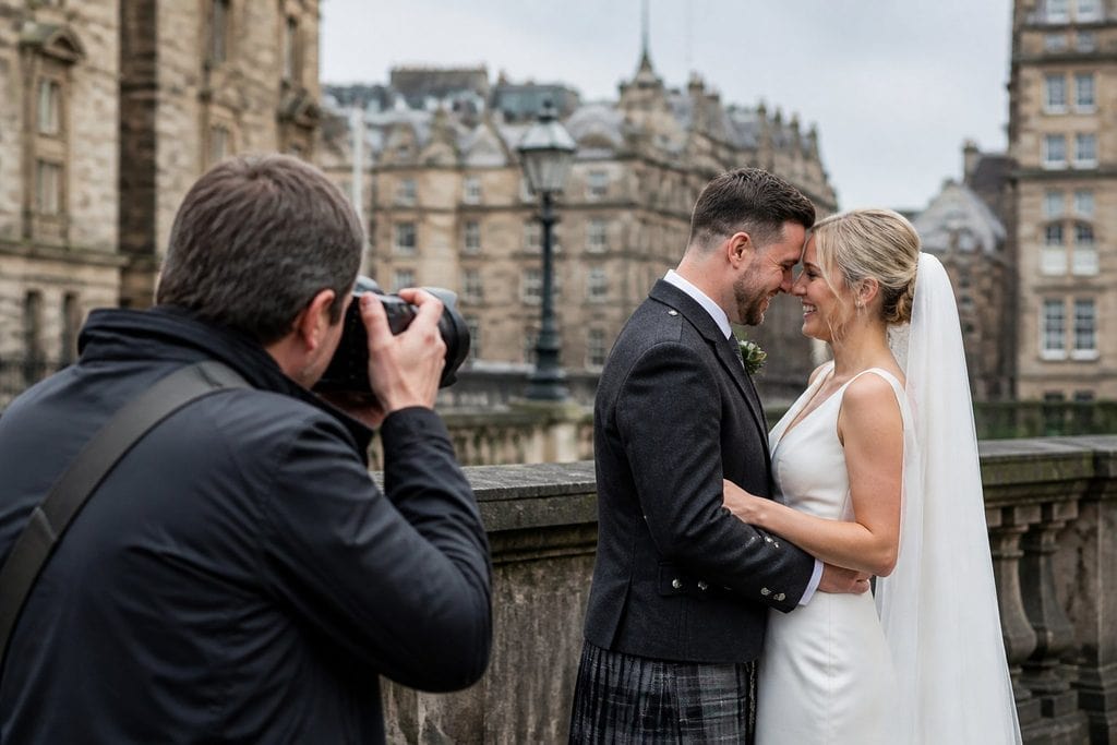 A photographer captures a bride and groom smiling and embracing outdoors in front of historic stone buildings on a cloudy day.